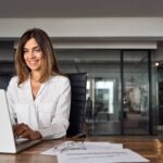 A smiling woman lawyer sits at a desk with a laptop in a real estate office, exuding professionalism and confidence.